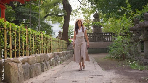 Slow motion of an Asian woman walking past a bamboo fence in a traditional Japanese garden. The path is lined with lush trees.