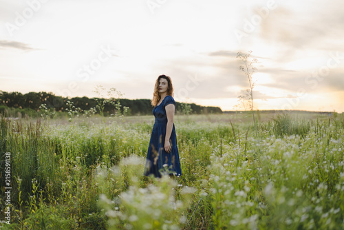 Canvastavla Young woman in a blue dress enjoying a peaceful summer evening in a blooming meadow at sunset