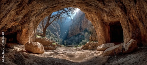 Panoramic view from inside a cave showing sunlight streaming through the opening, illuminating rocky terrain and a lush valley with trees in the background