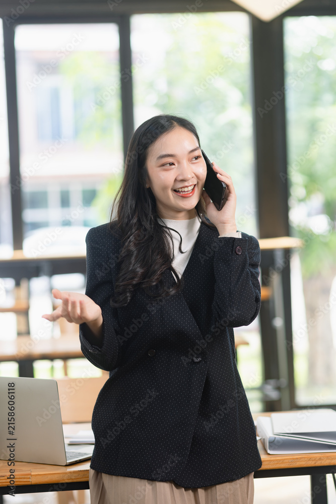 Fototapeta premium happy young Asian businesswoman talking on her smartphone and smiling while working in a modern office.