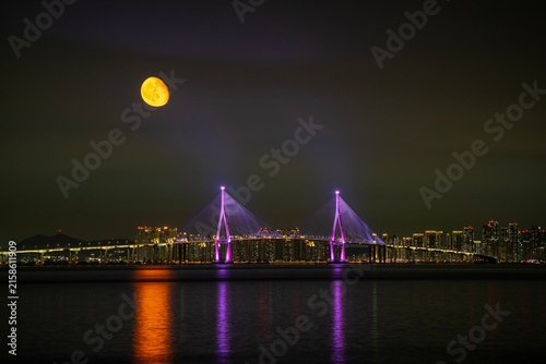 Full golden moon rising over illuminated Incheon Bridge at night with city skyline in the background, Incheon, South Korea.