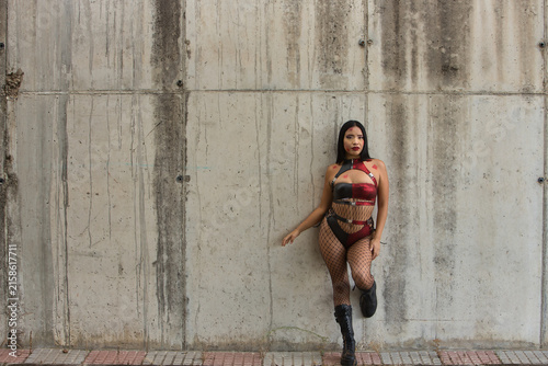 Young Latina woman, brunette, red eyes, wearing a leather suit and boots, fishnet stockings, posing leaning against a concrete wall in a post-apocalyptic city, as if she had become a zombie.