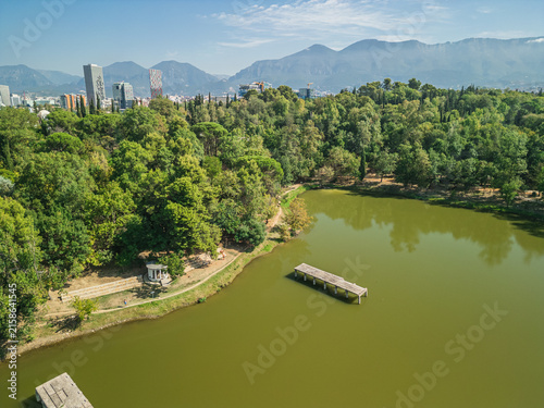 Photography Albania, Tirana 06 09 2025: Aerial drone photo of Grand Park lake with concrete platform surrounded by water, green trees, Marriott hotel, Downtown One and cloudy mountains