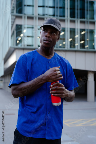 Картината върху платно Young black man in baseball jersey holding red water bottle, hydrating in urban