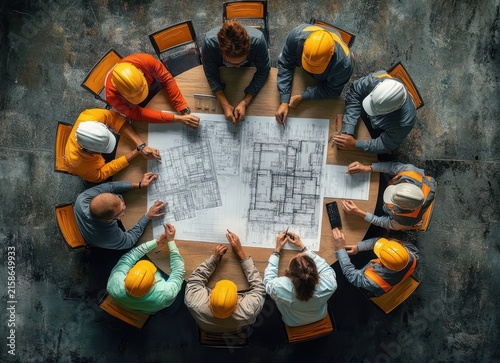 Overhead view of construction team in yellow and white hard hats and safety vests gathered around a table reviewing large blueprints, focused collaborative planning and discussion