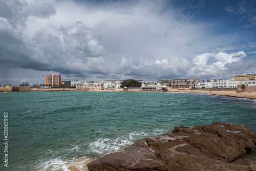 Photography Wide coastal view of La Caleta Beach in Cádiz, Spain, with turquoise waters, rocky foreground and dramatic clouds over the historic shoreline and seaside buildings