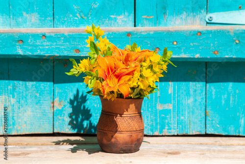 A bouquet of orange daylily and yellow loosestrife in a clay vase. Turquoise wooden background.