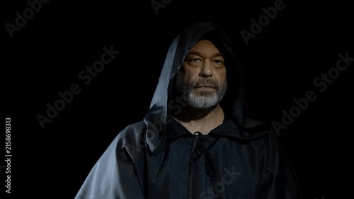 Hooded mature man slowly turns forward then lowers his gaze solemnly