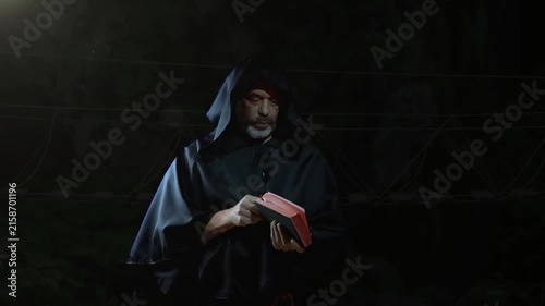 Hooded mature man slowly opens and reads red book in darkness