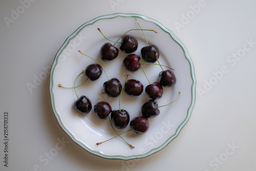 Foto Fresh dark sweet cherries with green stems arranged on a white ceramic plate, simple summer fruit composition on a light background with natural food styling