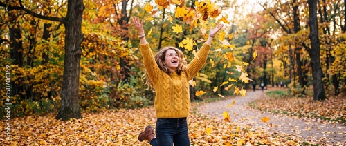Happy woman in yellow sweater throwing autumn maple leaves in air while jumping in forest park during sunny fall day with golden foliage