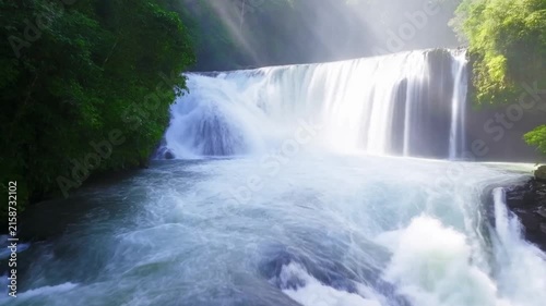 Powerful waterfall in lush tropical forest, water cascading over rocks, sun rays reflecting mist, serene natural landscape view.