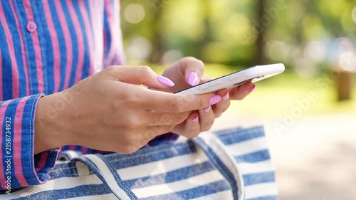 Close up of a woman with pink manicured nails using a white smartphone outdoors in a sunny park setting
