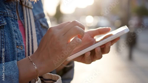 Close up of a woman typing and scrolling on her smartphone while standing outdoors in the city with warm sunlight.
