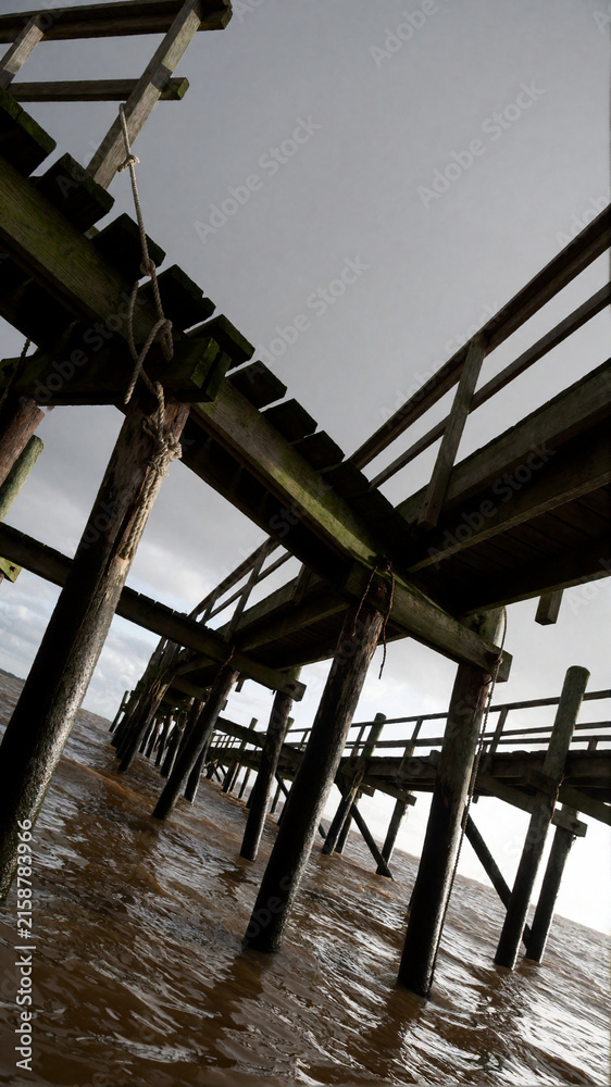 Fototapeta premium Low angle view of old wooden pier structure over muddy water under cloudy sky