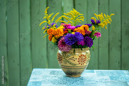 A bouquet of bright autumn flowers in a hand-painted ceramic vase against a green wooden wall.