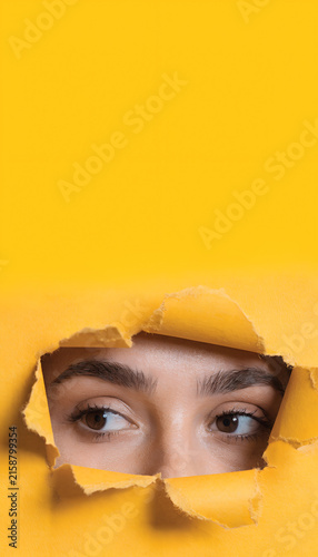 Close Up Of A Woman's Eyes Peeking Through A Torn Hole In Yellow Paper, Mysterious Curious Female Face Looking Through A Ripped Poster Background With Copy Space For Advertising And Promotion