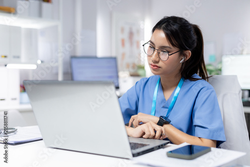 Healthcare professional in blue scrubs working on laptop in medical office, representing digital, patient records, clinical work, telehealth, nursing and medical technology.