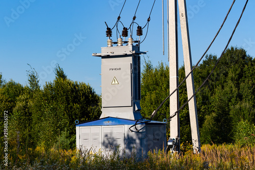 Compact electrical substation unit mounted on concrete poles for rural power supply, featuring high voltage transformers and grid cables amidst a green summer forest setting, 10 kilovolts