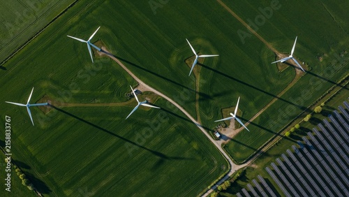 Top down aerial view of wind turbines casting long shadows across green agricultural fields with adjacent solar panel farm, sustainable renewable energy infrastructure generating clean energy from ab