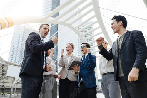 Group of business professionals celebrating a successful deal outdoors in a modern city, raising fists and smiling under contemporary architecture.