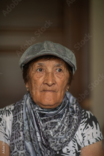 Fotografía An elderly woman with expressive dark eyes, wearing a gray newsboy cap and a patterned scarf, looking thoughtfully into the camera against a soft, blurred background