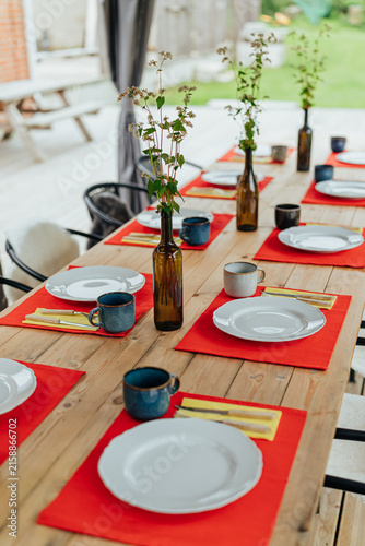 Canvastavla Long wooden table arranged with white plates, ceramic mugs, red placemats, cutle