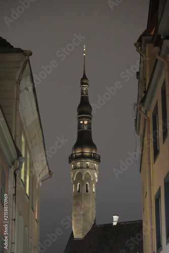 framed view of Tallinn Town Hall tower spire between historic alley buildings at night.