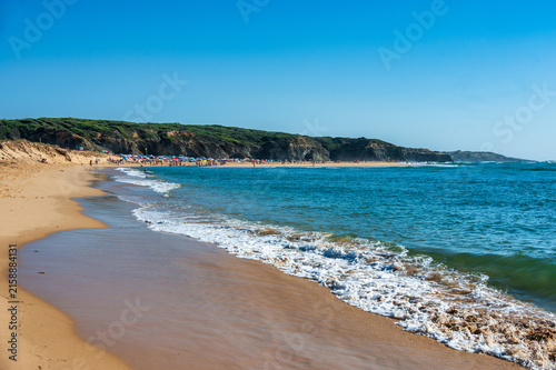 Waves rolling onto a golden sand beach with colorful umbrellas and cliffs