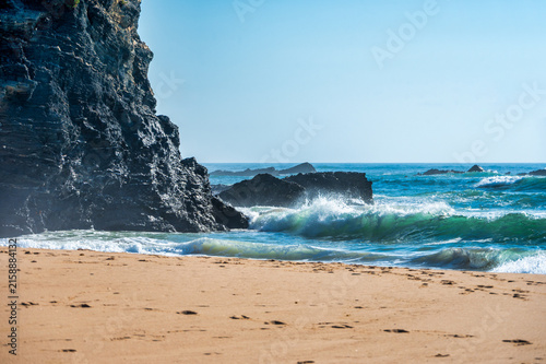 Turquoise ocean waves crash against dramatic dark rocky cliffs on a golden sand beach with footprints, under a clear blue sky, along the rugged Costa Vicentina coastline in the Alentejo, Portugal.
