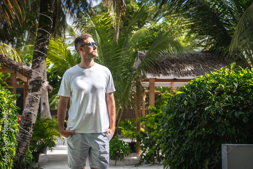 Photography A man wearing sunglasses and casual attire stands in a palm garden walkway at a tropical resort in Dhigurah island, Maldives, looking into the distance