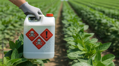 Hand holding a white container with hazard symbols in a green agricultural field glove 