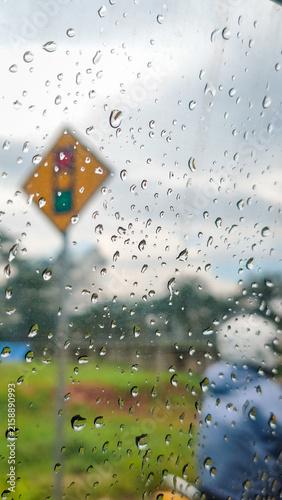 Raindrops on the car window with an out-of-focus red traffic light sign ahead. Crisp water droplets cluster on a windowpane while a yellow signal sign blurs softly in the wet background.
