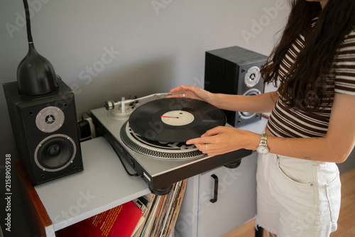 A person holds a vinyl record above a turntable in a room with audio speakers and a shelf of records nearby
