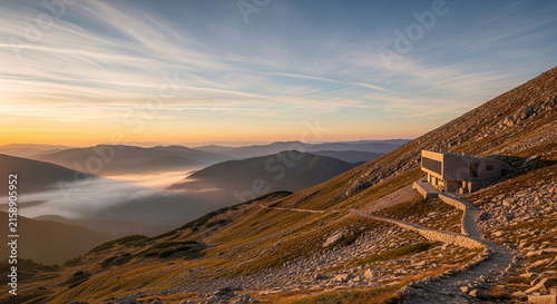 A contemporary wooden building sits on a steep mountain ridge overlooking a valley filled with fog during a golden hour sunset.