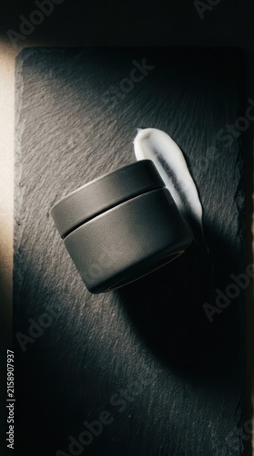 Cosmetic jar rests on a dark slate tile next to a smear of white moisturizing lotion.