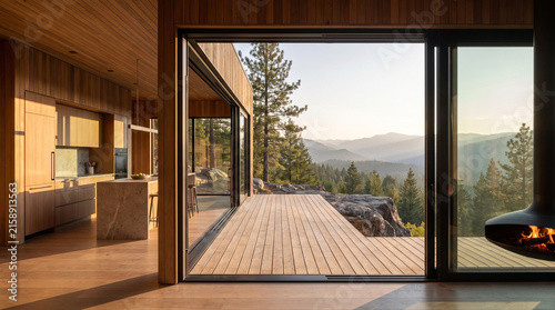 Interior view of a minimalist wooden house featuring a marble kitchen island and a terrace with mountain forest views at sunset.