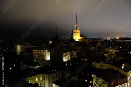 high-angle view of Tallinn Old Town at night, featuring medieval city wall defense towers and the illuminated St. Olaf's Church spire