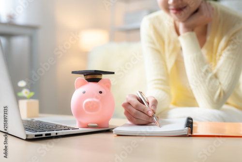 Obraz na płótnie Woman planning education savings with piggy bank in graduation cap, laptop and notebook at home, representing college fund, student finance, budgeting and future education goals