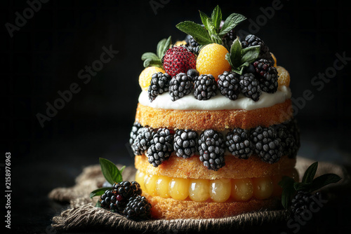 A cake with two visible layers features soft sponge and fresh fruits. Berries and herbs rest on top, adding color and texture. The scene is set in a kitchen with low light