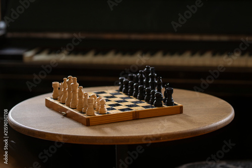 Obraz na płótnie Vintage wooden chessboard with black and white chess pieces arranged for a strategic game on a round wooden table, with a piano softly blurred in the background