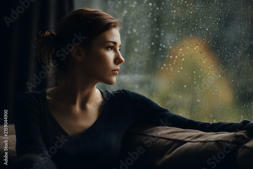 Woman sits alone on couch looking at rainy window