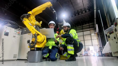 Collaboration with Robotics: Two engineers in safety vests and hard hats collaborate closely with a sophisticated robotic arm on a factory floor, reviewing plans and discussing its operation.