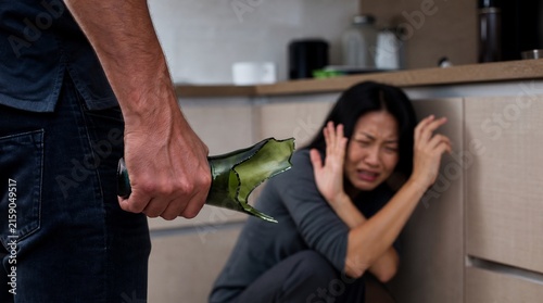 Aggressive Man Holding Broken Glass Bottle Threatening Cowering Woman in Kitchen, Concept of Domestic Violence and Spousal Abuse