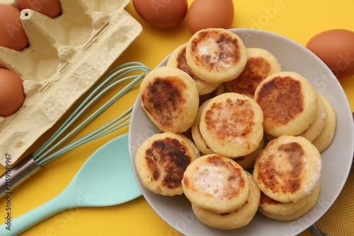 Homemade delicious syrniki, cheese pancakes in plate, cooking ingredients, kitchen utensils on yellow background
