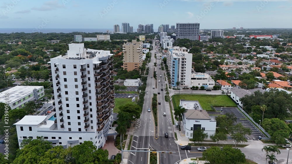 Fototapeta premium Aerial view of a modern white residential high rise tower beside a wide palm lined boulevard in a Miami neighborhood with the Atlantic Ocean on the horizon