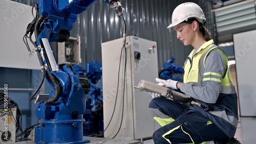 Industrial Robotics Technician: A young female technician in a hard hat and safety vest inspects a large industrial robot arm in a factory setting.