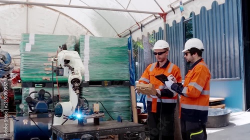 Robotic Arm Collaboration: Two engineers in protective gear observe a robotic arm in action within an industrial setting, discussing progress and technology.