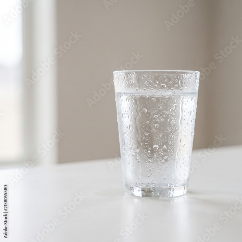 Glass of Water on White Table: A close-up shot of a clear glass filled with cold water, condensation forming droplets on the exterior, sitting on a clean white surface.