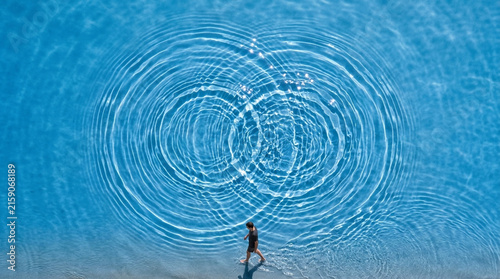 High angle view of clear blue water with overlapping circular ripples creating an infinity pattern, with a person walking at the bottom edge.